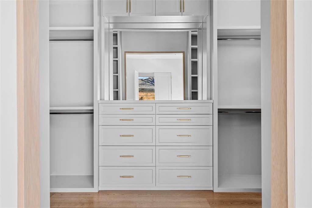 This walk-in closet boasts custom-built cabinetry with ample storage, elegantly accented by chic brass cabinet pulls. A clerestory window allows natural light to filter in, enhancing the airy ambiance. The rich white oak hardwood floors add warmth and sophistication, creating a beautifully organized and inviting dressing space.