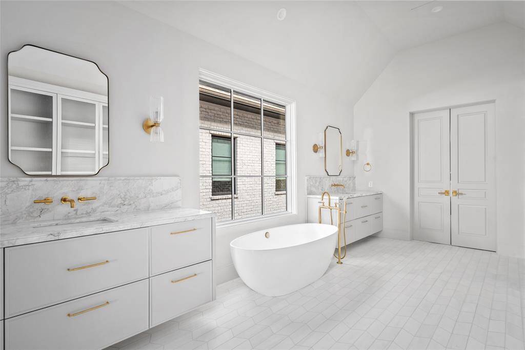 Exuding timeless elegance, the custom-designed primary bathroom features marble dual vanities lit by Aerin sconces. A freestanding soaking tub with a striking brass tub filler sits beneath a large window, enhancing the airy ambiance.