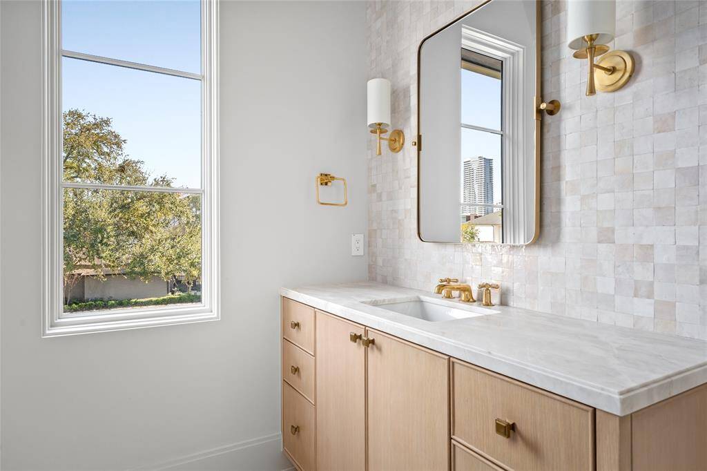 This secondary bathroom features a custom vanity with marble counters, accented by honey bronze cabinet hardware and walls adorned with Zellige tile, which complement the Aerin sconces that provide a warm glow. A shower/tub combo is finished with handmade artisan tile walls and is paired with a brass shower kit. Underfoot, limestone tile floors add a timeless touch.