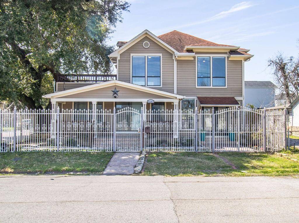 This charming two-story home features a classic design with large windows, a welcoming porch, and a decorative metal fence. The property is surrounded by mature trees, offering ample shade and privacy.