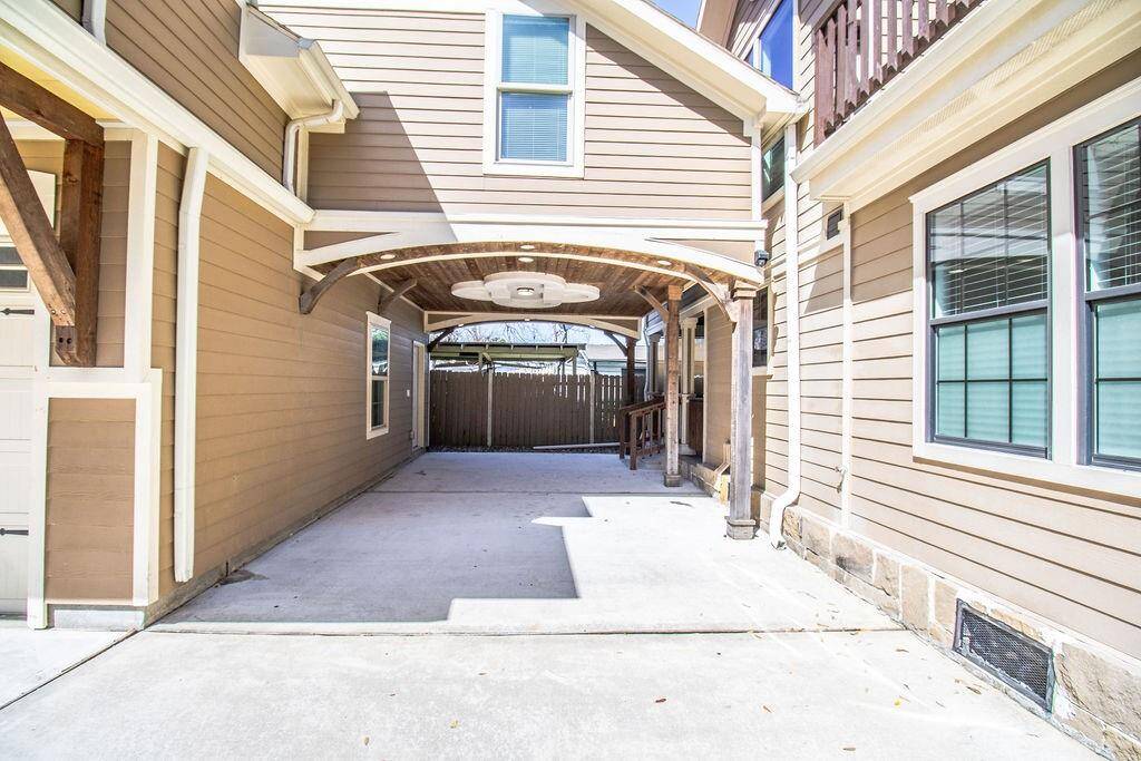 This photo showcases a covered driveway or carport area with a wooden ceiling and recessed lighting, nestled between two sections of a house. The exterior features beige siding and white trim, offering a clean, modern look.
