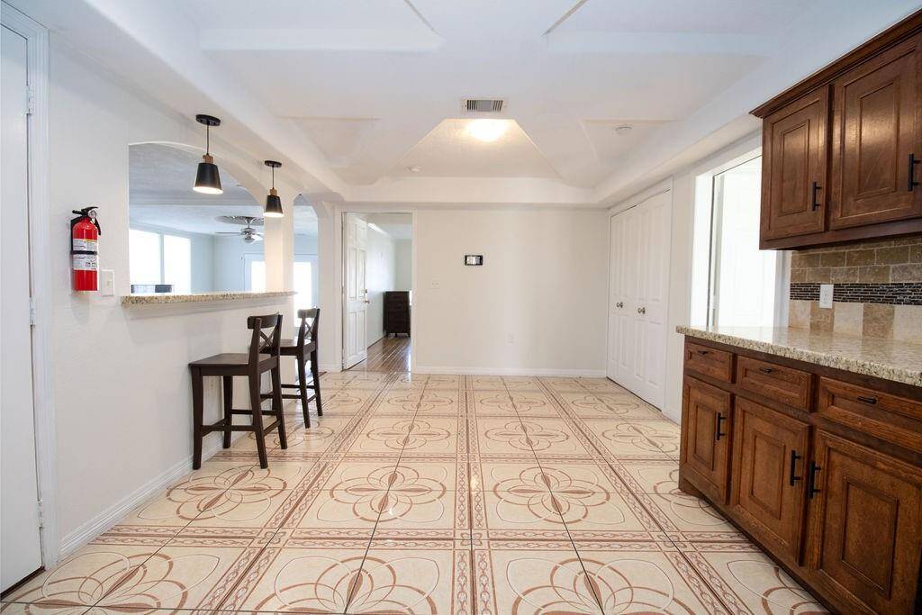 This spacious kitchen area with decorative tiled flooring and a bar area with pendant lighting and stools. It boasts ample natural light, wooden cabinets with granite countertops, and leads into an open living space.