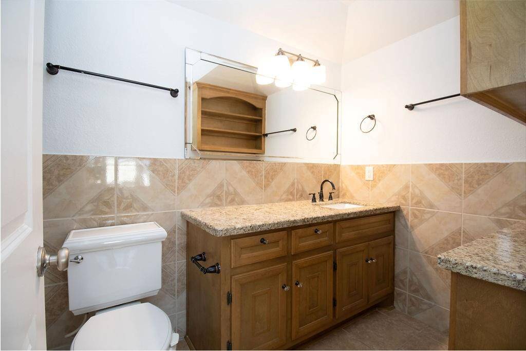 This master bathroom features a granite countertop with a wooden vanity, a wall-mounted mirror, and modern lighting. It has tiled walls and flooring, a clean white toilet, and ample storage space.
