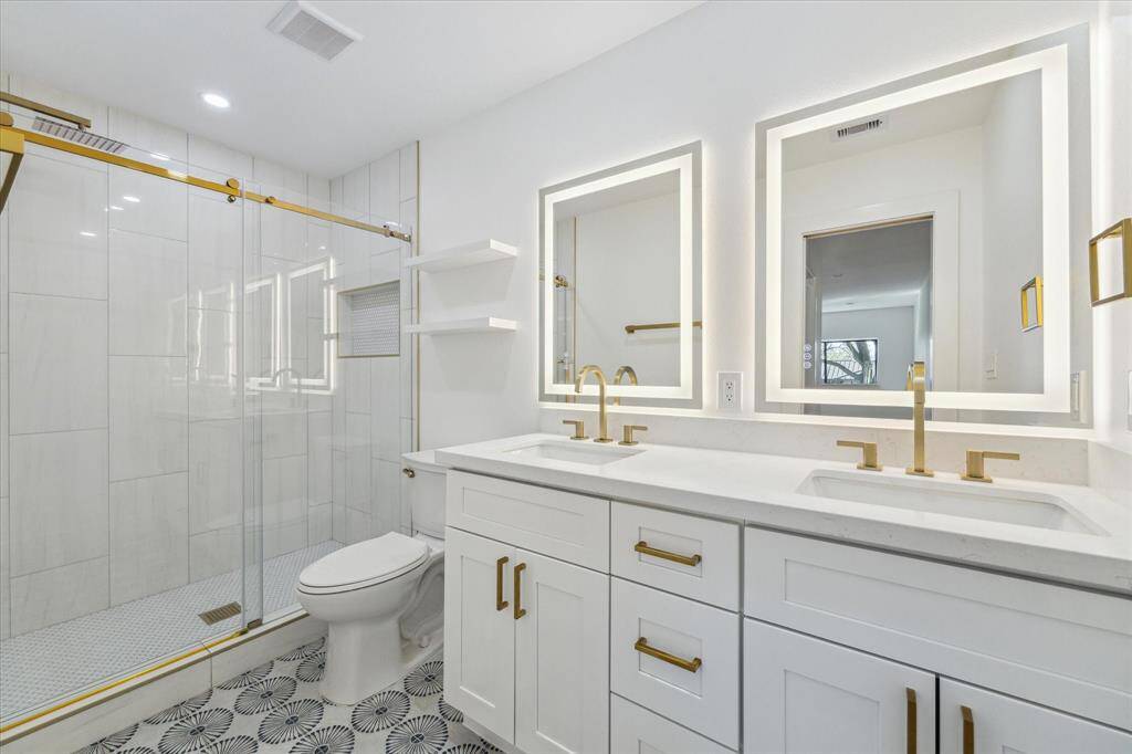 This modern primary bathroom features a spacious glass-enclosed shower with elegant gold fixtures, dual sinks with illuminated mirrors, and stylish patterned floor tiles, creating a luxurious and contemporary feel.