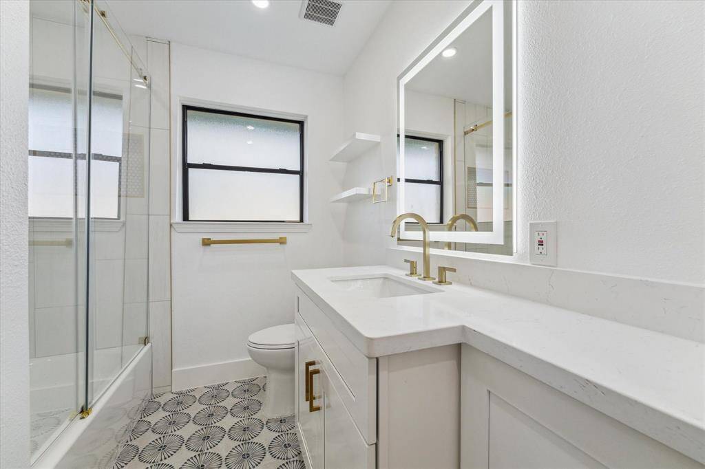 The hall bathroom features a modern design with a tub and shower combo, elegant patterned floor tiles, and a large illuminated mirror. The vanity has sleek gold fixtures and a clean, white countertop. Frosted windows provide natural light and privacy. Stackable washer and dryer connections located in hall bath.