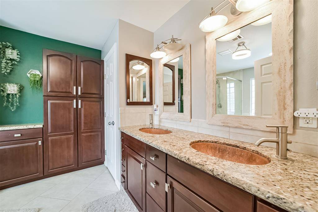 The bathroom features a copper hammerred double-sink vanity with granite countertops and stylish lighting. It includes ample dark wood cabinetry for storage and a vibrant green accent wall with decorative plants, creating a fresh and inviting space.