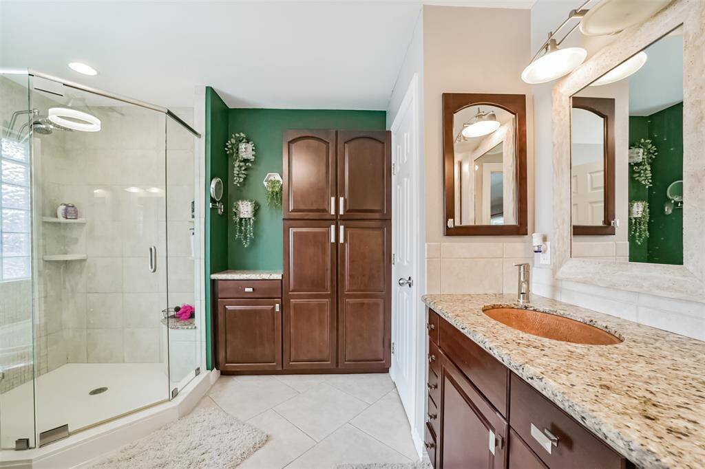 This modern bathroom features a double-sink vanity with granite countertops and stylish lighting. It includes ample dark wood cabinetry for storage and a vibrant green accent wall with decorative plants, creating a fresh and inviting space.