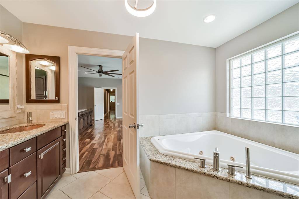 This bathroom features a spacious corner jacuzzi tub with a sleek granite surround, a modern vanity with dual sinks and elegant fixtures, plus ample natural light from a frosted glass window. The room flows into a hallway with warm wood flooring and a ceiling fan, offering a seamless transition to the rest of the home.