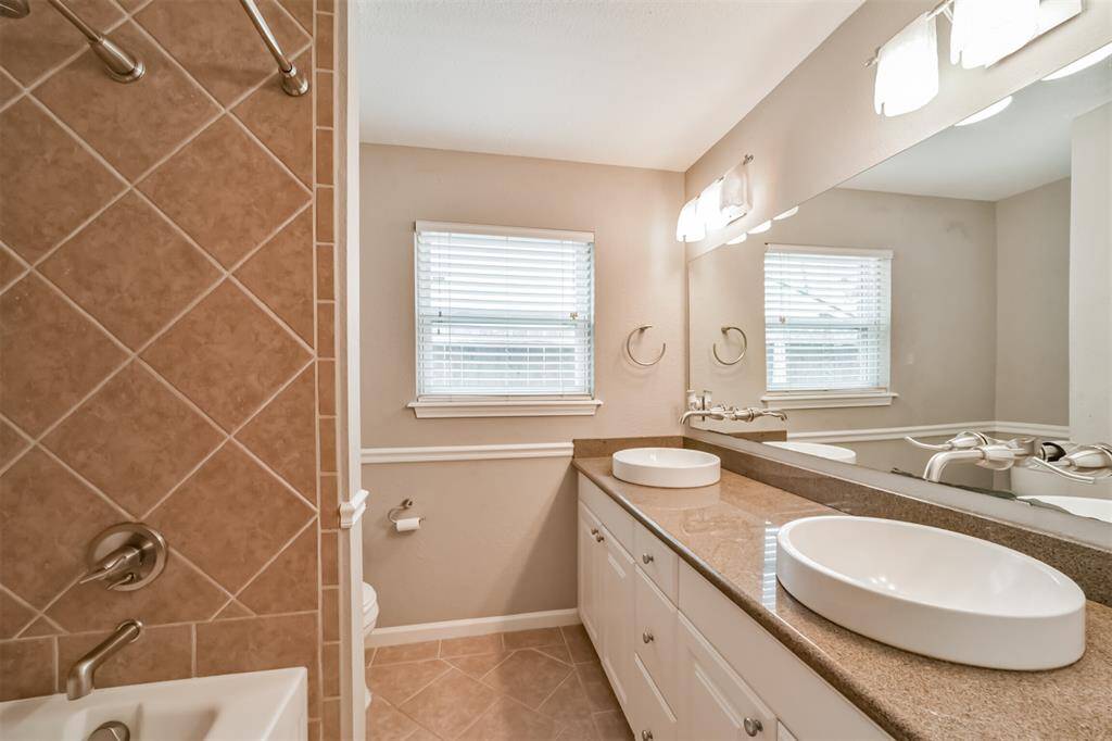 This bathroom features a modern design with dual vessel sinks on a spacious granite countertop. It includes a tiled shower and bath area, ample lighting, and two windows for natural light.