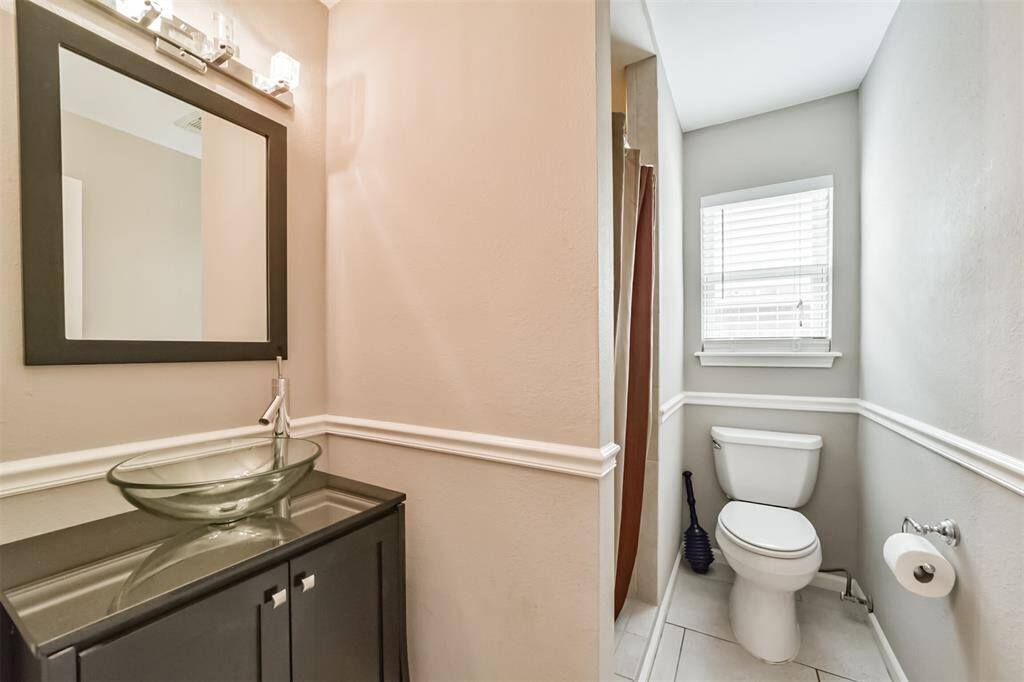 This bathroom features a modern glass vessel sink with a sleek faucet, under a large framed mirror. It includes a separate toilet area with natural light from a window, and subtle wall detailing adds a touch of elegance.
