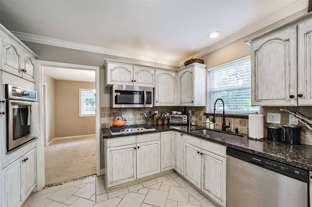 his kitchen features rustic cabinets, a modern granite countertop, and a stainless steel sink with a sleek faucet. The backsplash has a warm, earthy tile design, and there's a window providing natural light.
