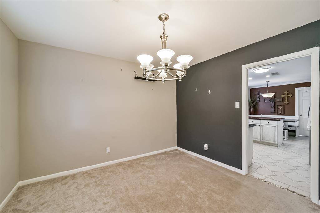 This photo showcases a small dining or living space with neutral carpet and walls, accented by a dark feature wall. A modern chandelier provides overhead lighting. The room connects to an adjacent kitchen area with white cabinetry and tile flooring.