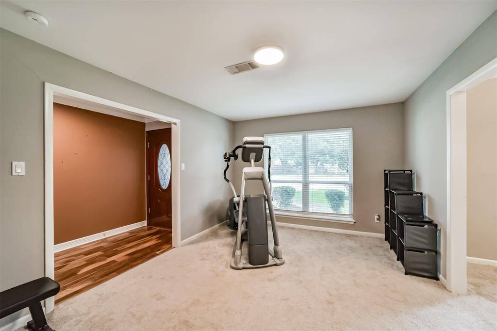 This room features neutral walls and carpet, creating a cozy atmosphere. It includes a large window with ample natural light, perfect for a home gym or additional living space. The room connects to an area with warm wood flooring and a stylish front door.