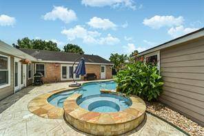 This photo showcases a spacious backyard with a stone-edged swimming pool and attached spa, surrounded by a concrete patio. The area includes a covered seating space, ideal for outdoor relaxation and entertainment. The yard is enclosed by a privacy fence, enhancing the home's appeal and providing a serene outdoor setting.