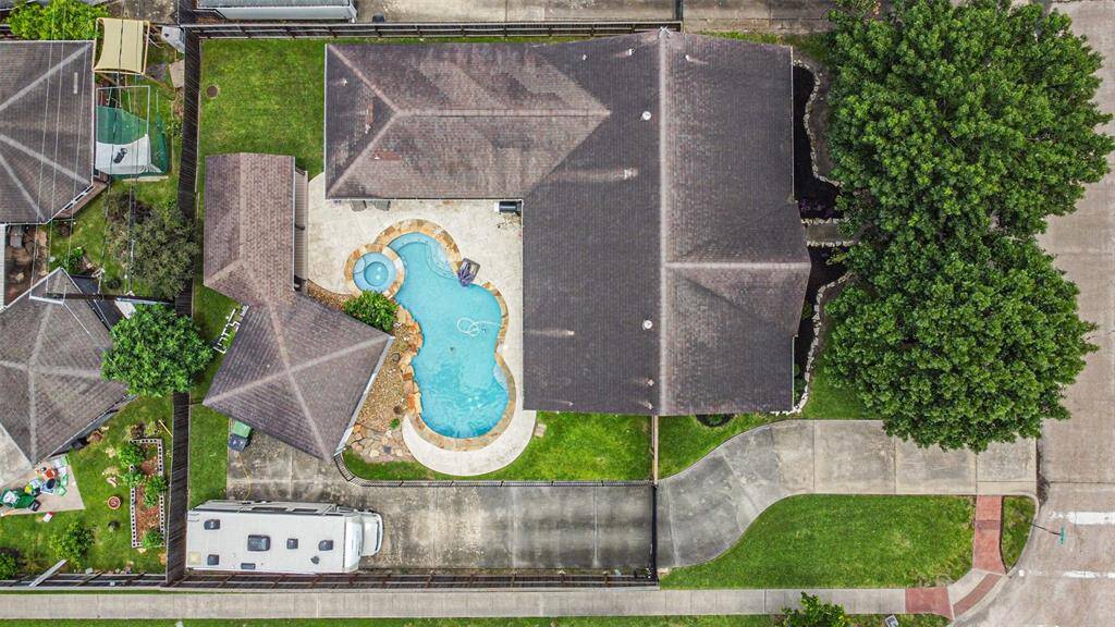 This aerial view showcases a spacious home with a uniquely shaped pool and spa, surrounded by a well-maintained lawn. The property includes a driveway and is bordered by trees, offering a mix of privacy and open space.