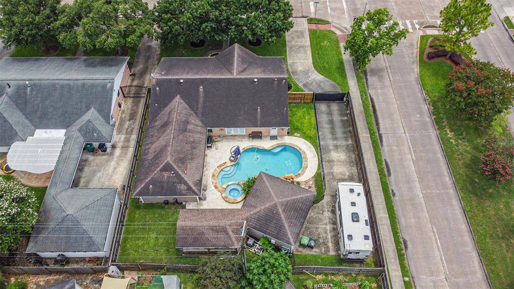 This aerial view shows a spacious property featuring a central swimming pool with a surrounding patio. The house has a large, pitched roof and an adjacent grassy area. There is a driveway with additional parking, including space for an RV. The property is bordered by trees and situated on a corner lot.