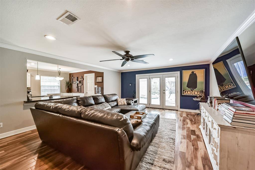 This inviting living room features a spacious layout with a large sectional sofa, wood flooring, and a ceiling fan. It opens to the kitchen, creating a seamless flow. A wall-mounted TV and cozy decor complete the space.