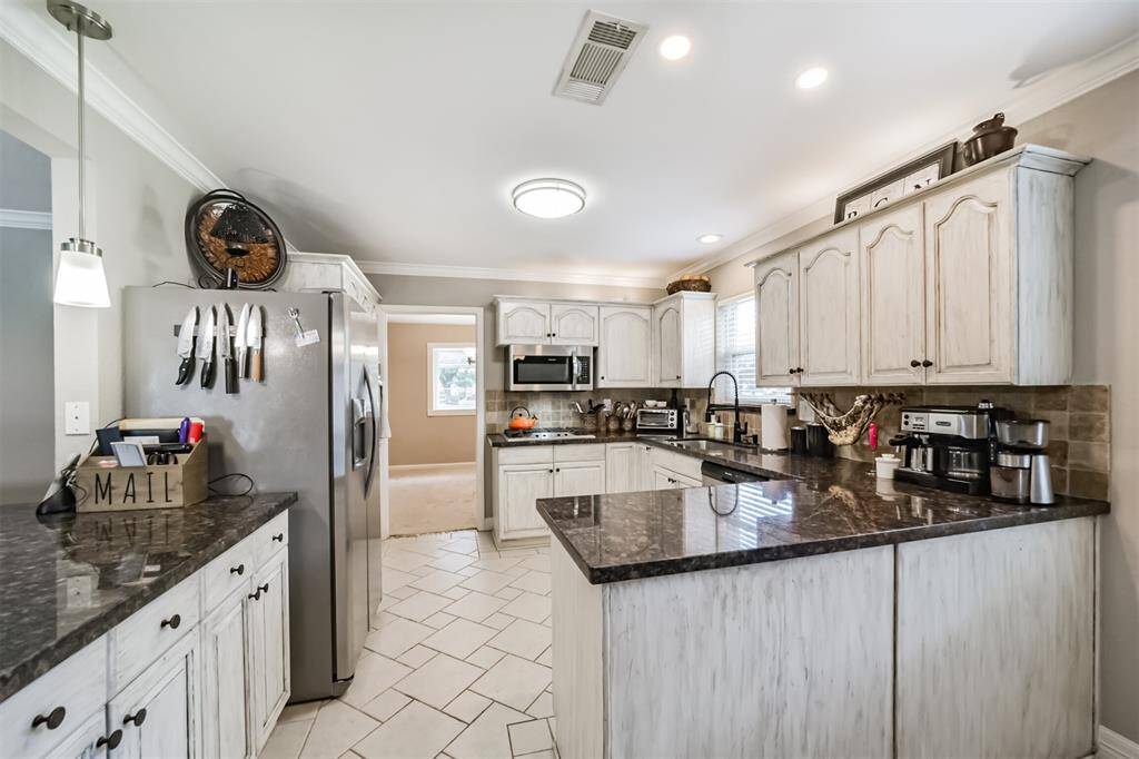 This kitchen features rustic white cabinetry with dark countertops, stainless steel appliances, and a cozy tile backsplash. A large window provides natural light, and there's a spacious layout with a visible adjacent room.