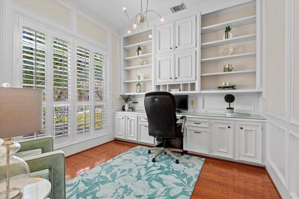 This is a bright home office space featuring white built-in cabinetry and shelving, large windows with plantation shutters, and elegant hardwood floors.