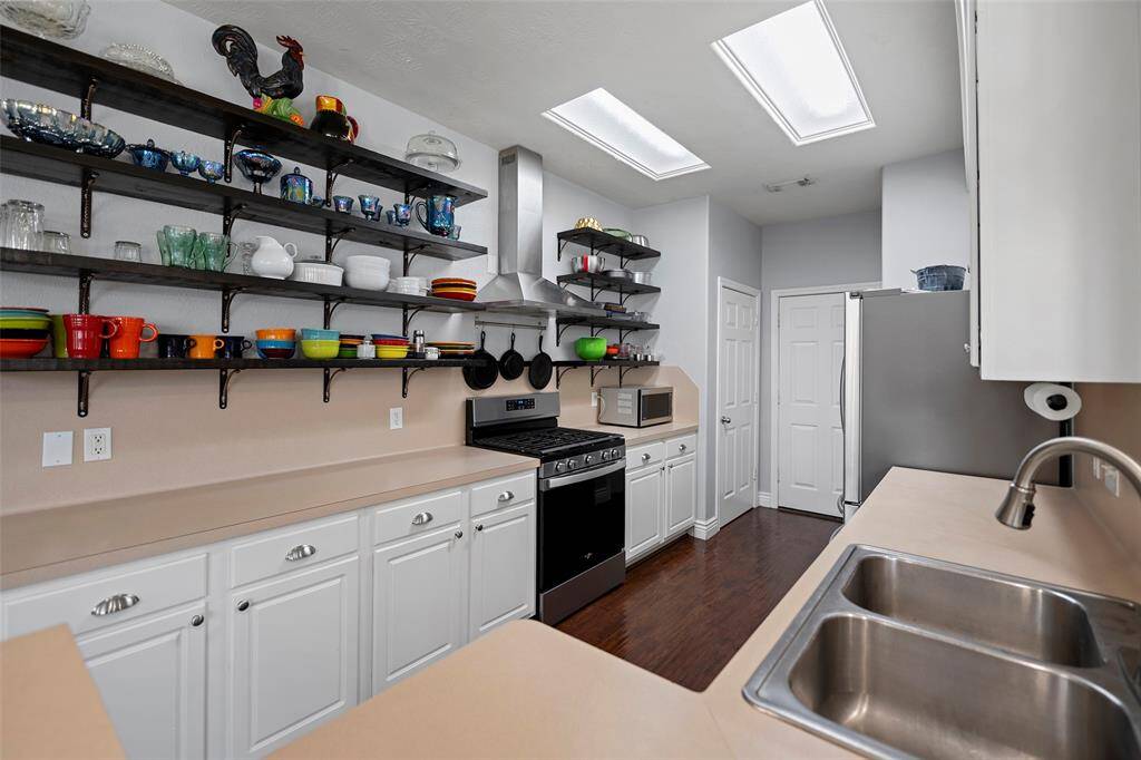 This kitchen features white cabinetry with open shelving for display. The space is well-lit by overhead skylights, and the dark wood flooring adds warmth and contrast.