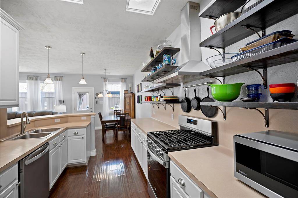 This bright kitchen features modern open shelving, stainless steel appliances, and a spacious layout with hardwood floors. It opens to a dining area, creating a seamless flow for entertaining.