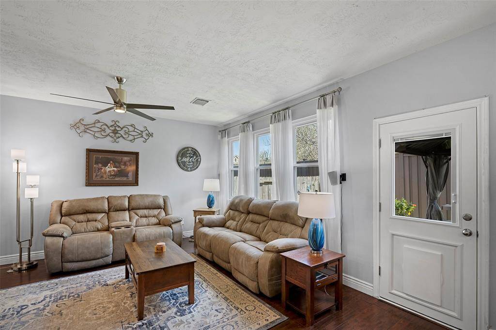 This inviting living room features a modern ceiling fan, and large windows with sheer curtains providing natural light with the convenient side door leading outside.