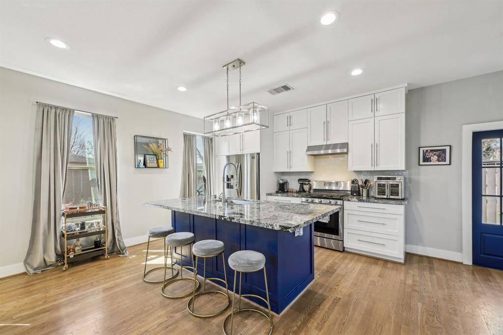 This photo showcases a modern kitchen with white cabinetry, stainless steel appliances, and a large granite island with seating. The space is open and bright, featuring hardwood floors and ample natural light from adjacent windows.