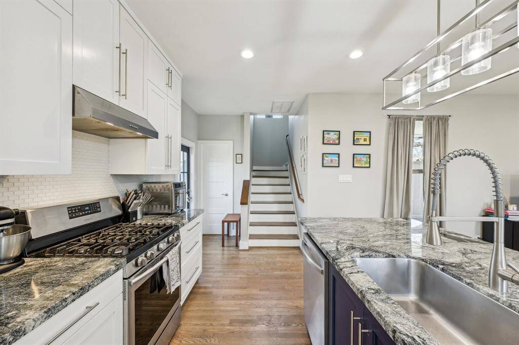 This photo shows a modern kitchen with white cabinets and stainless steel appliances. It features a gas range, granite countertops, and a large fridge. Natural light streams in from a window with curtains, creating a bright and inviting space. A framed picture adds a personal touch to the wall.