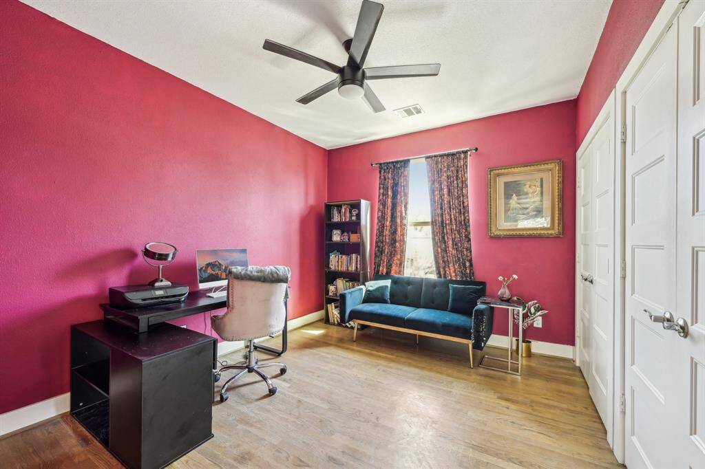 This bedroom features vibrant magenta walls, a stylish ceiling fan, and hardwood floors. It includes a cozy bed with an upholstered headboard, ample natural light from a window, and access to a walk-in closet.