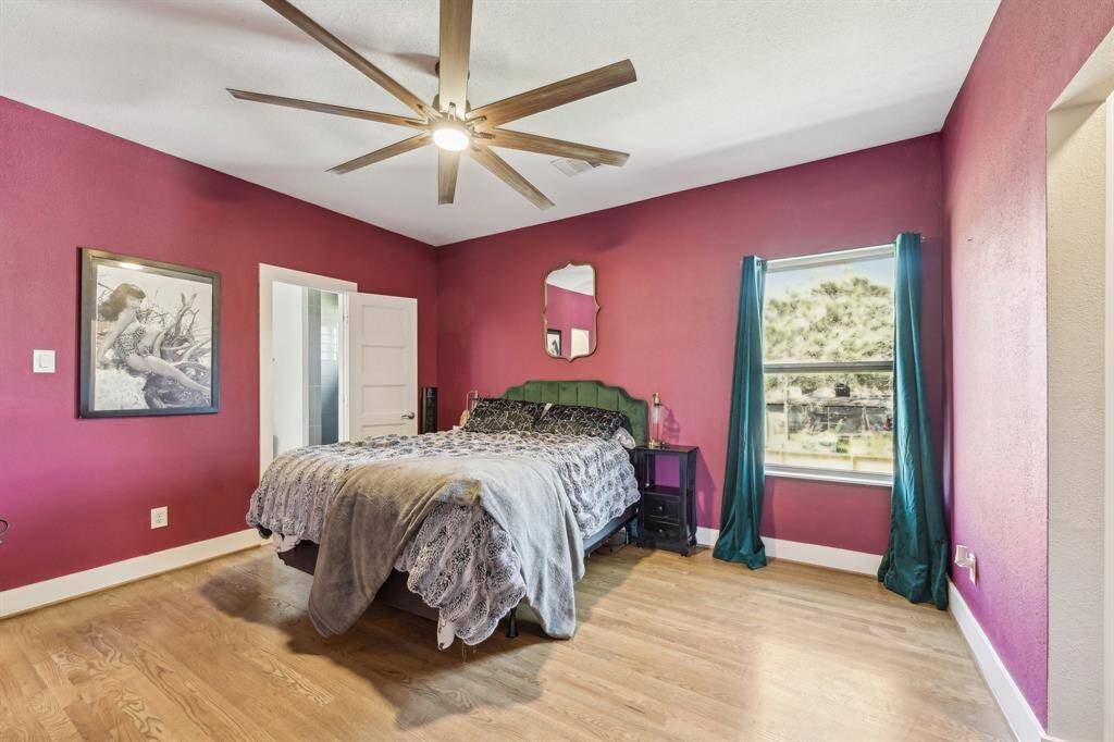 This bedroom features vibrant magenta walls, hardwood floors, and a large ceiling fan. It offers direct access to an adjoining room and a bathroom, and includes modern decor with a mirror and dresser.