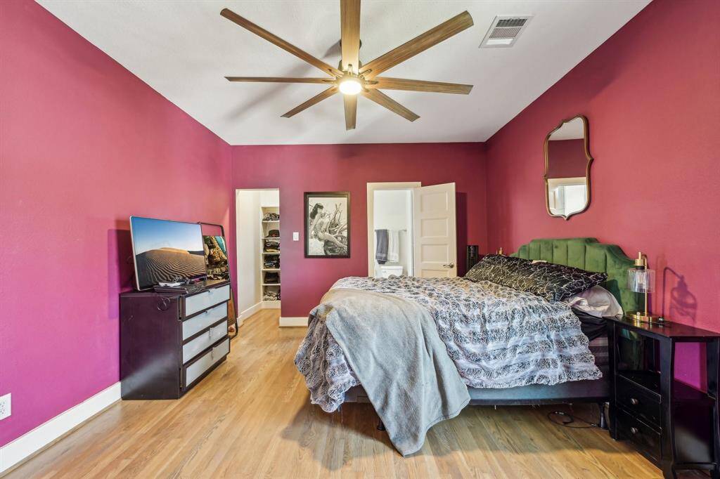This spacious bedroom features vibrant red walls and wood flooring. It includes a ceiling fan, large bed, and modern furnishings. The room connects to a walk-in closet and a bathroom, offering both comfort and convenience.