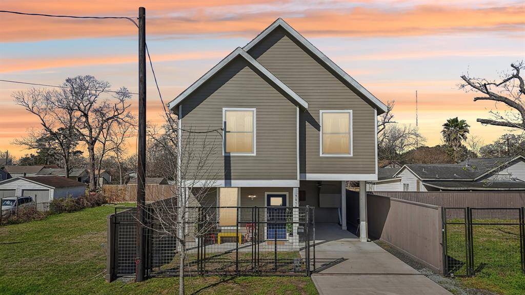 This modern two-story home features a sleek exterior with a gabled roof and a unique elevated design. It includes a gated driveway, a spacious front yard, and a welcoming porch. The setting is serene, with mature trees and a vibrant sunset sky in the background.