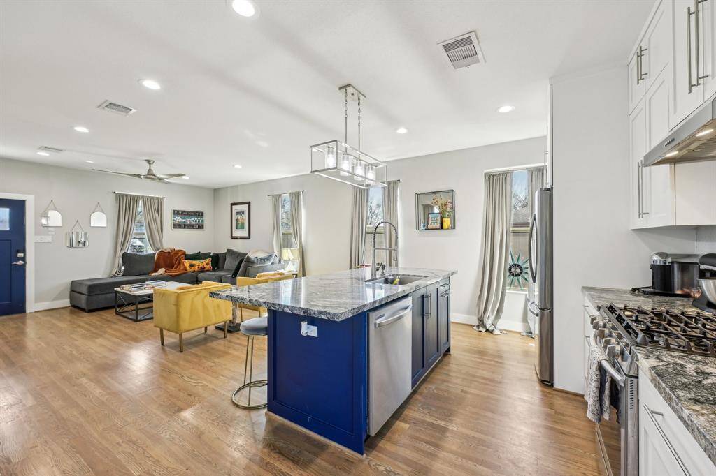 Bright and inviting living room with hardwood floors, a modern ceiling fan, and ample natural light from multiple windows. Features a cozy gray sectional, mustard armchairs, and a bold blue front door for a pop of color.