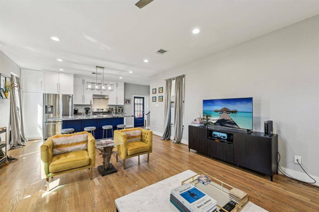 Bright and inviting living room featuring hardwood floors, neutral walls, and recessed lighting. The room includes a cozy gray sectional, two mustard-colored chairs, a flat-screen TV, and a blue front door. Natural light streams in through the windows with light curtains, creating a warm atmosphere.