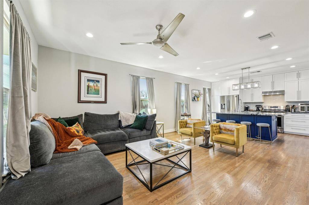 This modern kitchen features a spacious island with a blue base and marble countertop, ideal for casual dining with four stools. It boasts stainless steel appliances, white cabinetry, and hardwood floors, complemented by large windows with elegant curtains for ample natural light.
