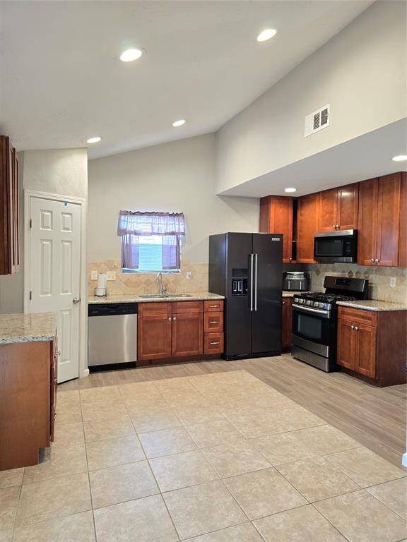 Kitchen features stainless steel appliances, warm wood cabinetry, and a spacious layout with tile flooring. Recessed lighting and a window above the sink provide ample illumination.