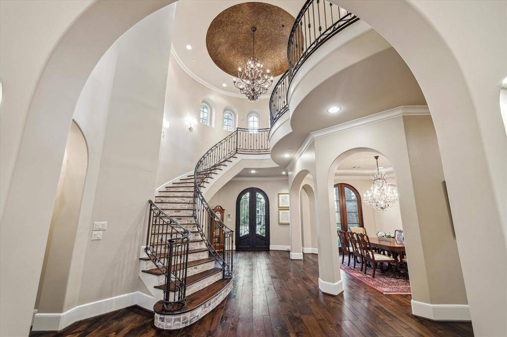 Elegant entry foyer featuring a dramatic curved staircase has iron railings. The high ceiling is crowned with a large, round golden dome that enhances the space's luxurious feel complemented by a sparkling chandelier.