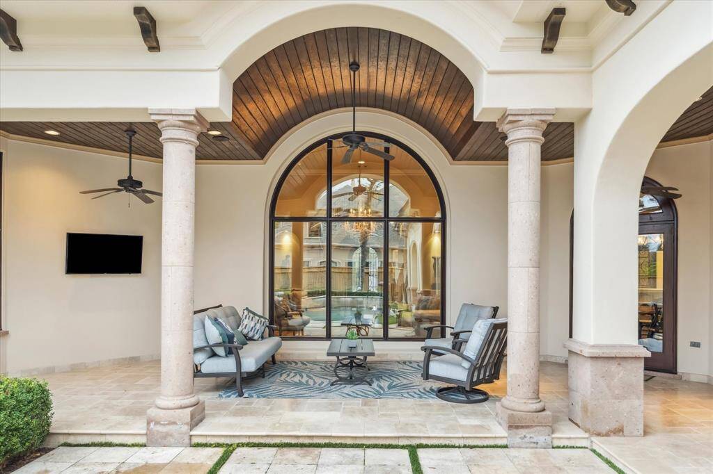 The covered patio boasts impressive stone columns, elegant curved archways, creating a luxurious and welcoming atmosphere. The ceiling is adorned with rich wood beading and equipped with ceiling fans and TV.