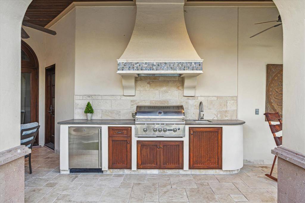 Summer kitchen with outdoor grill, sink, small fridge, and custom hood adding an exquisite design element.
