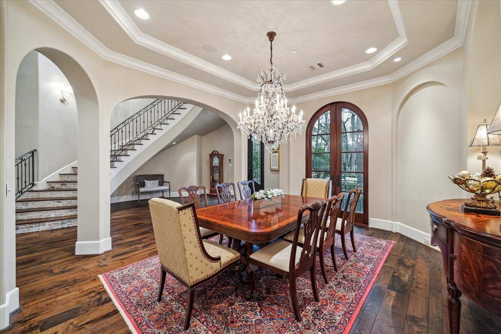 A formal dining room featuring crown molding, arched doorways, and a luxurious chandelier. Tall double French doors with arches allow natural light to fill the room.