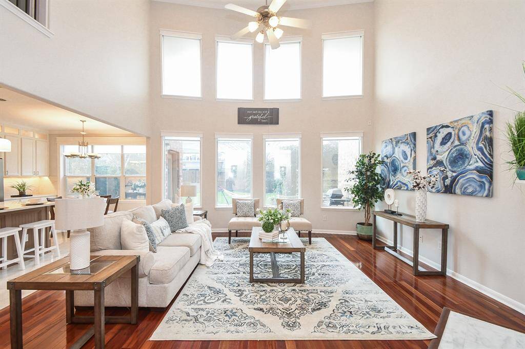 This spacious living room features high ceilings and abundant natural light from large windows. It includes elegant wood flooring, a cozy seating area, and tasteful decor. The open layout connects seamlessly to the kitchen and dining area, ideal for entertaining. The lower windows have remote operated shades.