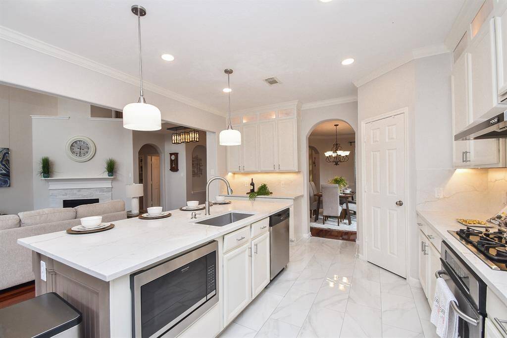 This modern kitchen features sleek white cabinetry, stainless steel appliances, and a spacious island with bar seating. The open layout flows into a cozy living area with a fireplace and elegant dining space, creating a welcoming atmosphere for entertaining.