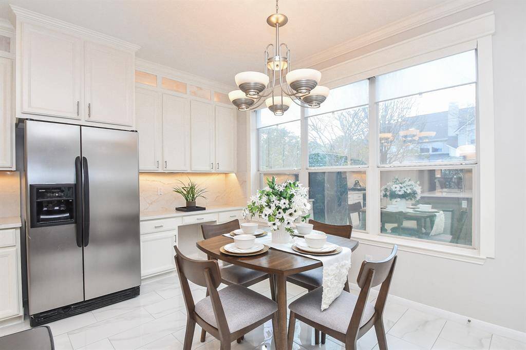This bright kitchen features modern stainless steel appliances, ample white cabinetry, and a cozy dining area with a round table set for four. Large windows provide plenty of natural light, complemented by a stylish chandelier.