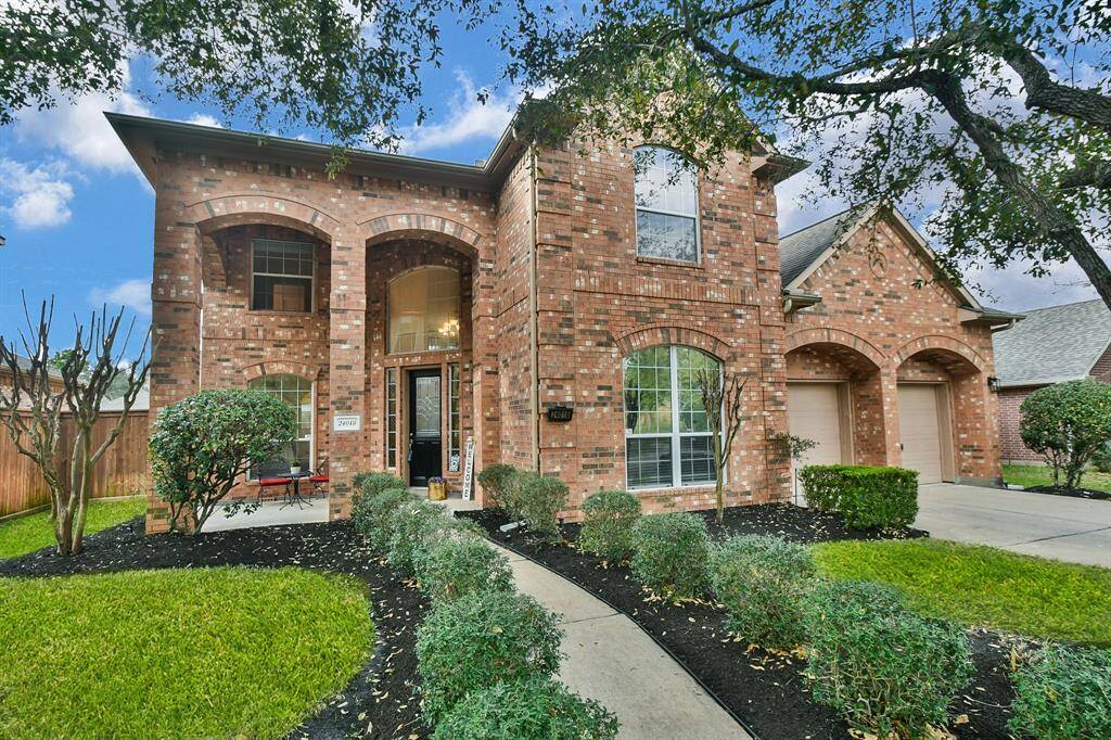This charming two-story brick home features a welcoming entrance with arched windows and a well-maintained front yard. It includes a double garage and a curved walkway leading to the front door, surrounded by lush greenery.