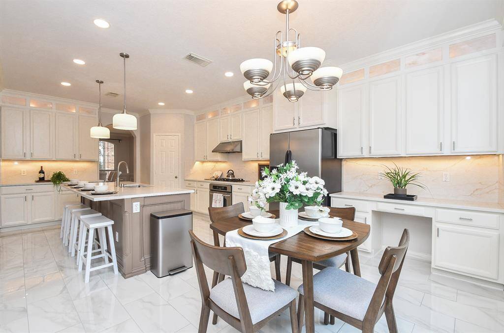 This photo showcases a spacious, modern kitchen with sleek white cabinetry, a large island with bar stools, stainless steel appliances, and elegant lighting fixtures. The room is bright with ample counter space and a cozy dining area.