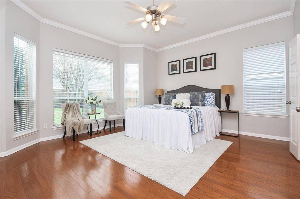 This bright and spacious bedroom features large windows with abundant natural light, hardwood floors, and neutral walls. It includes a cozy sitting area, a ceiling fan, and elegant decor, offering a comfortable and inviting atmosphere.