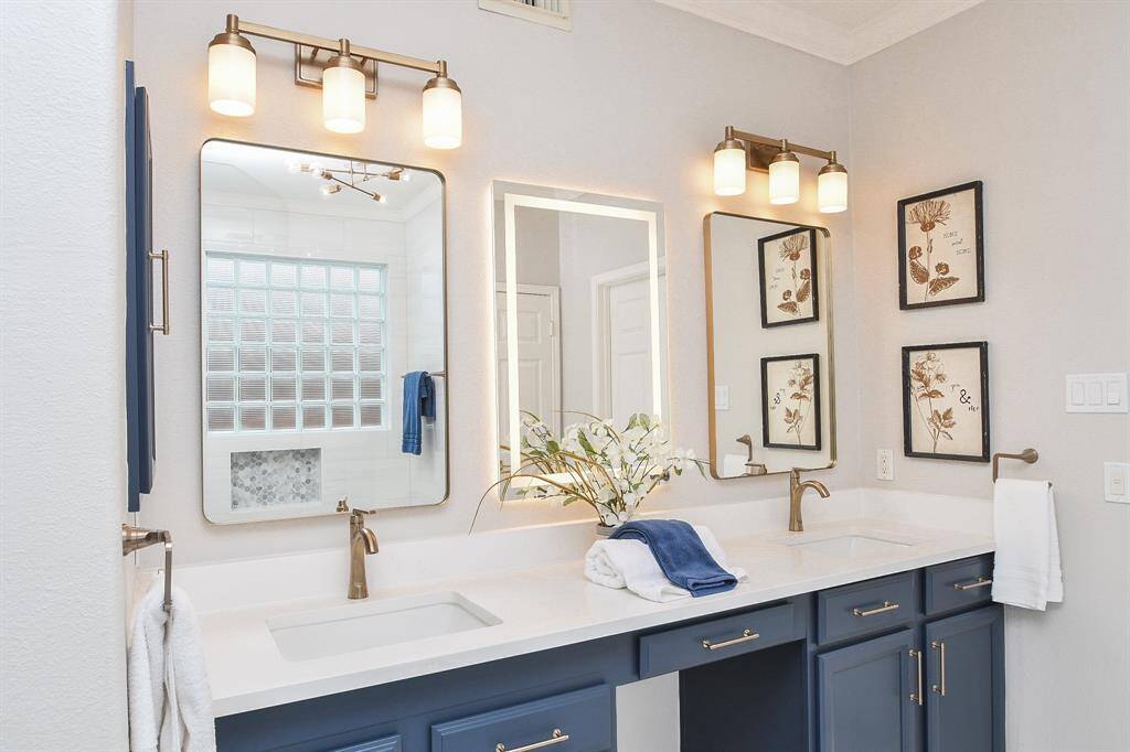This bathroom features a modern dual vanity with sleek white quartz countertops and freshly painted cabinets. It includes elegant bronze fixtures, large mirrors, and ample lighting. All the light fixtures, mirrors, and faucets have been replaced recently.