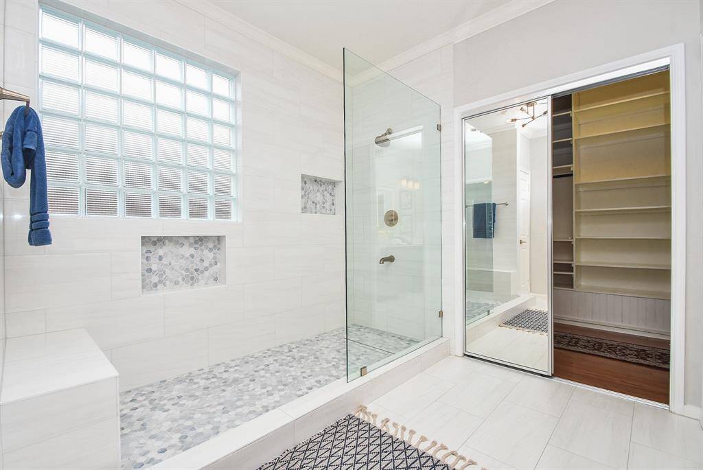 This bathroom features a spacious glass-enclosed shower with stylish tile work and built-in niches. It offers ample natural light through a glass block window and includes a sliding mirror door leading to a walk-in closet with shelving.
