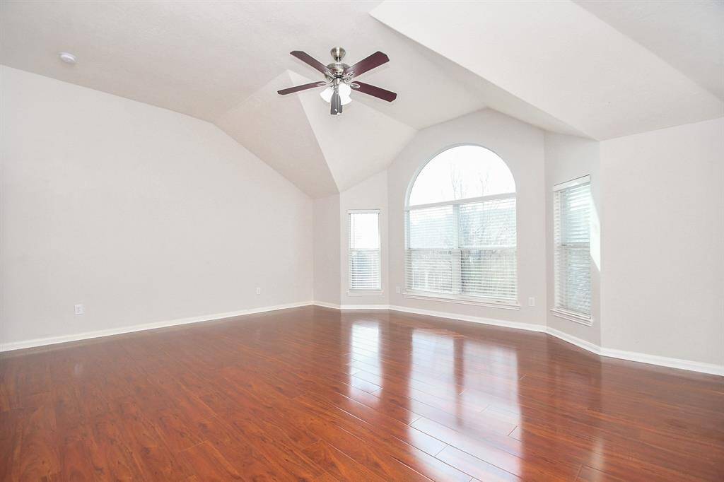 This spacious game room features high vaulted ceilings and rich wood flooring, complemented by a large, arched window that provides ample natural light. A ceiling fan adds functionality and style.