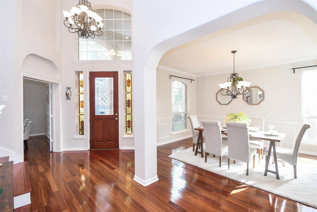 This inviting entryway features a rich wood door with elegant stained-glass panels, leading into a bright dining area with large windows. The space boasts high ceilings, beautiful hardwood floors, and classic chandeliers, creating a warm and sophisticated atmosphere.